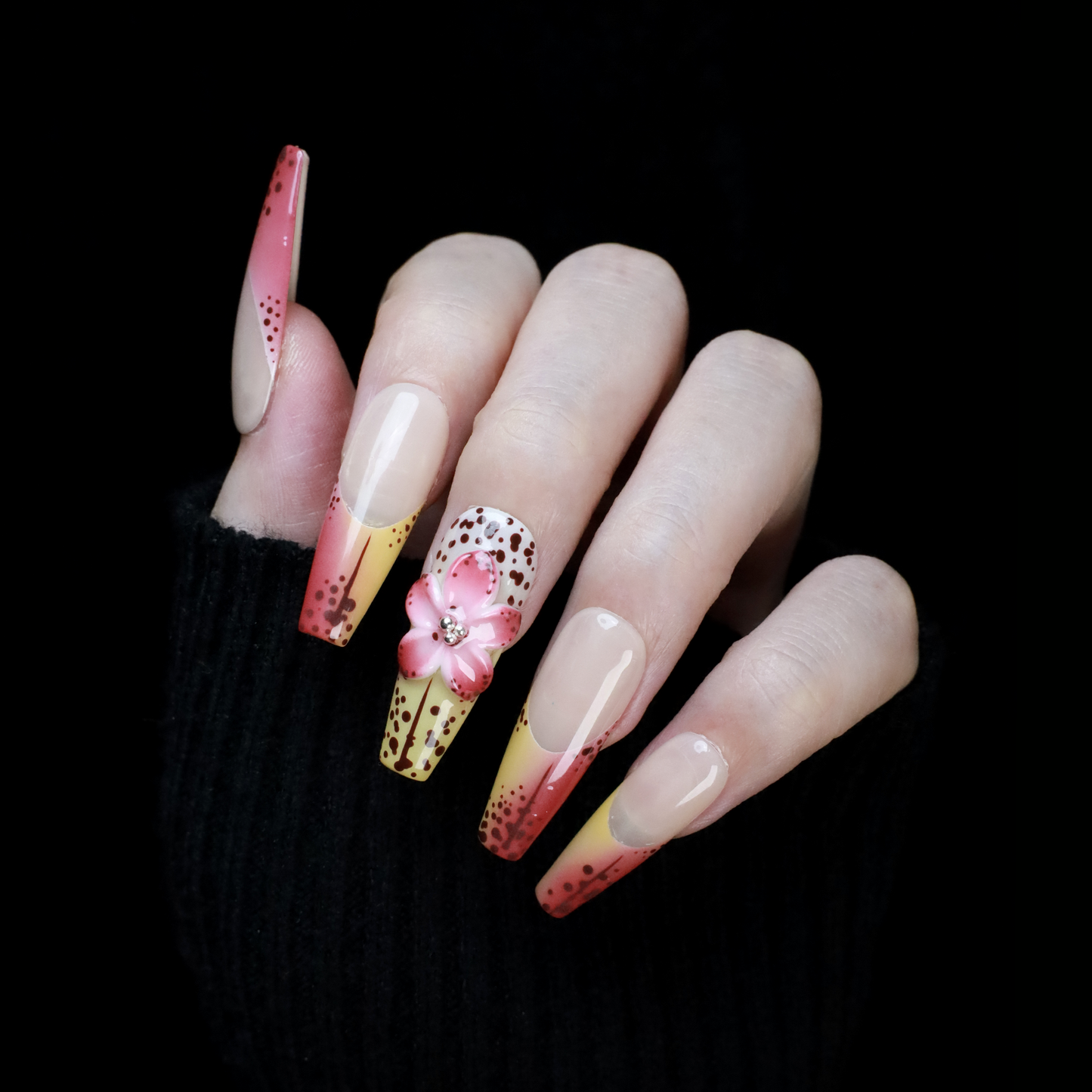 A hand showcases We Do Nails Sunset Bloom Press-On Nails featuring a pink-to-yellow gradient with black dots. One nail displays a 3D pink flower with a pearl on a black background, perfectly paired with a sleek sweater sleeve.