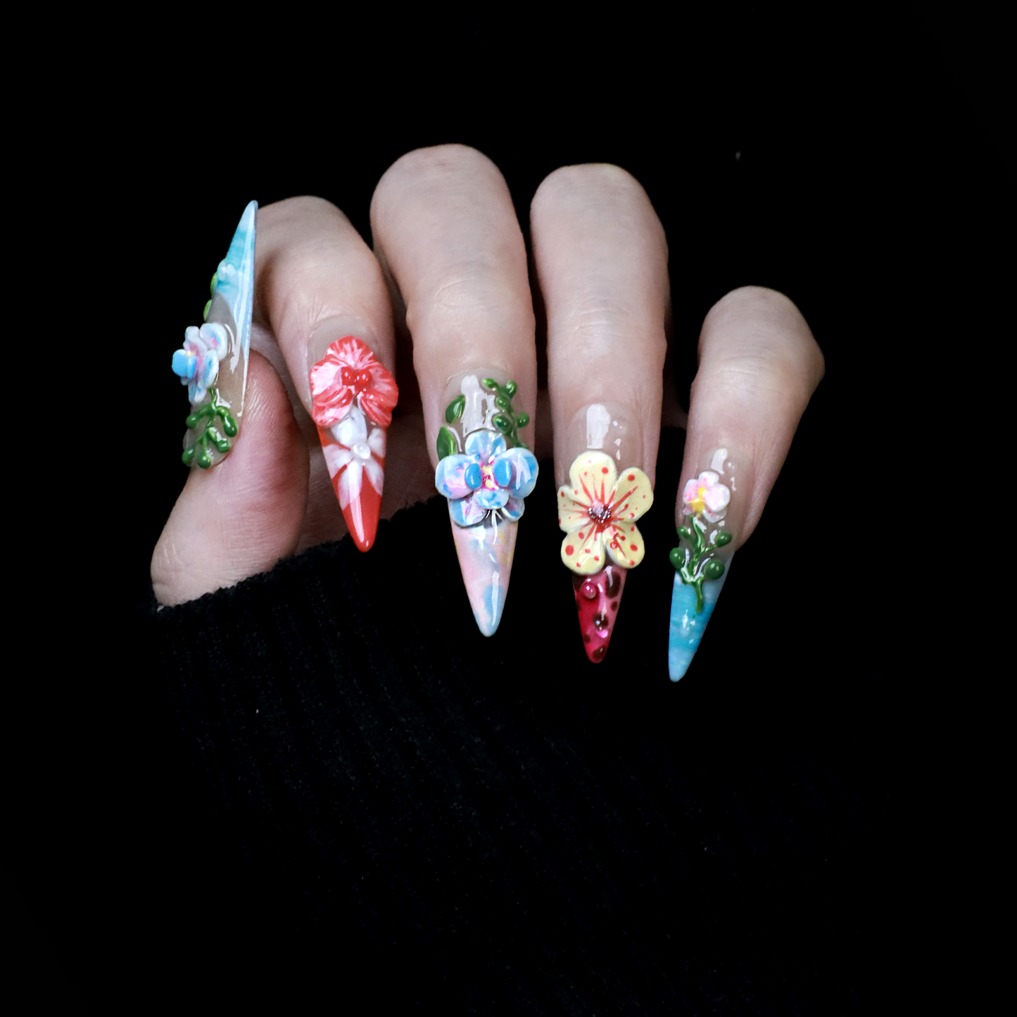 A hand shows off We Do Nails Floral Paradise Press-On Nails with long, pointed acrylics adorned with 3D flowers in shades of red, pink, yellow, and green. Set against a dark background, the vibrant blossoms truly stand out.