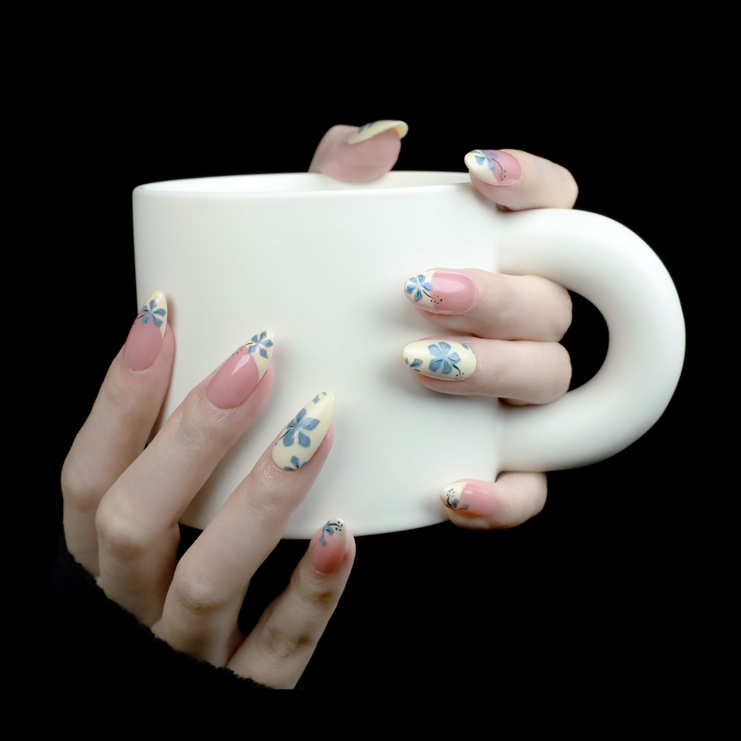 Elegant hands sporting We Do Nails Blue and White Porcelain Elegance Press On Nails hold a large white mug against a black background.