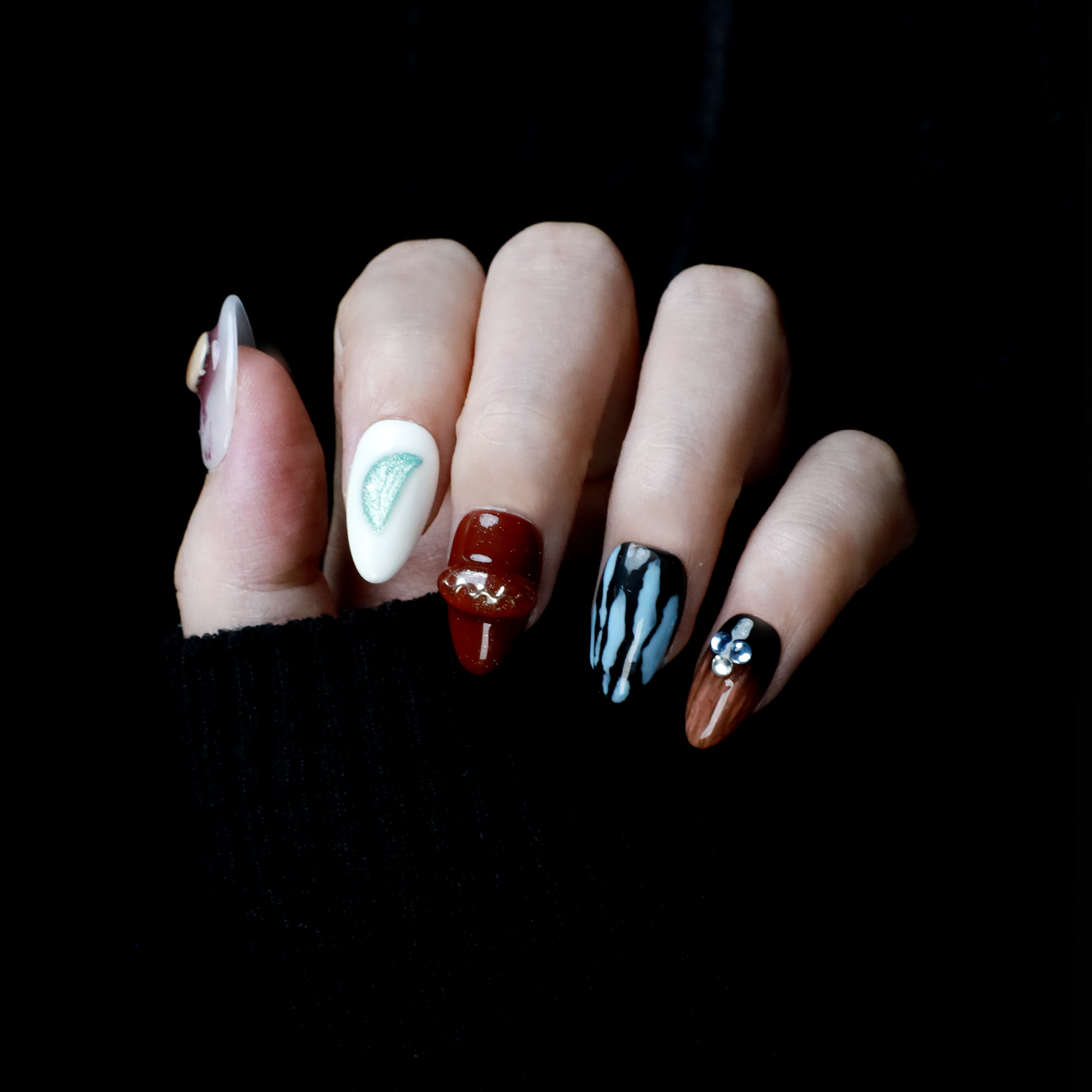 A hand elegantly displays Avant-Garde Luxe Press-On Nails by We Do Nails against a black backdrop. These nails, featuring 3D embellishments, boast white, maroon, and black hues with intricate patterns and metallic accents.