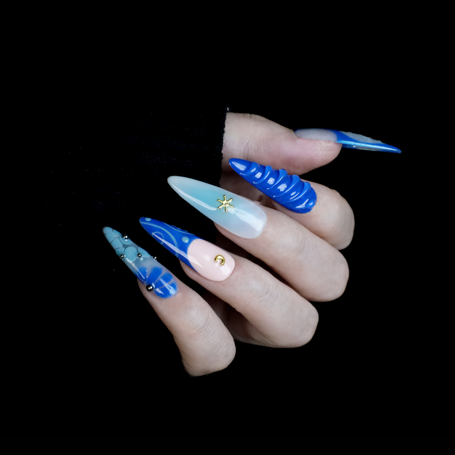 A hand featuring We Do Nails Blue Dreams Press-On Nails with elongated designs and pink hues, includes 3D metallic nail art and textured patterns like ridges and dots against a black backdrop.