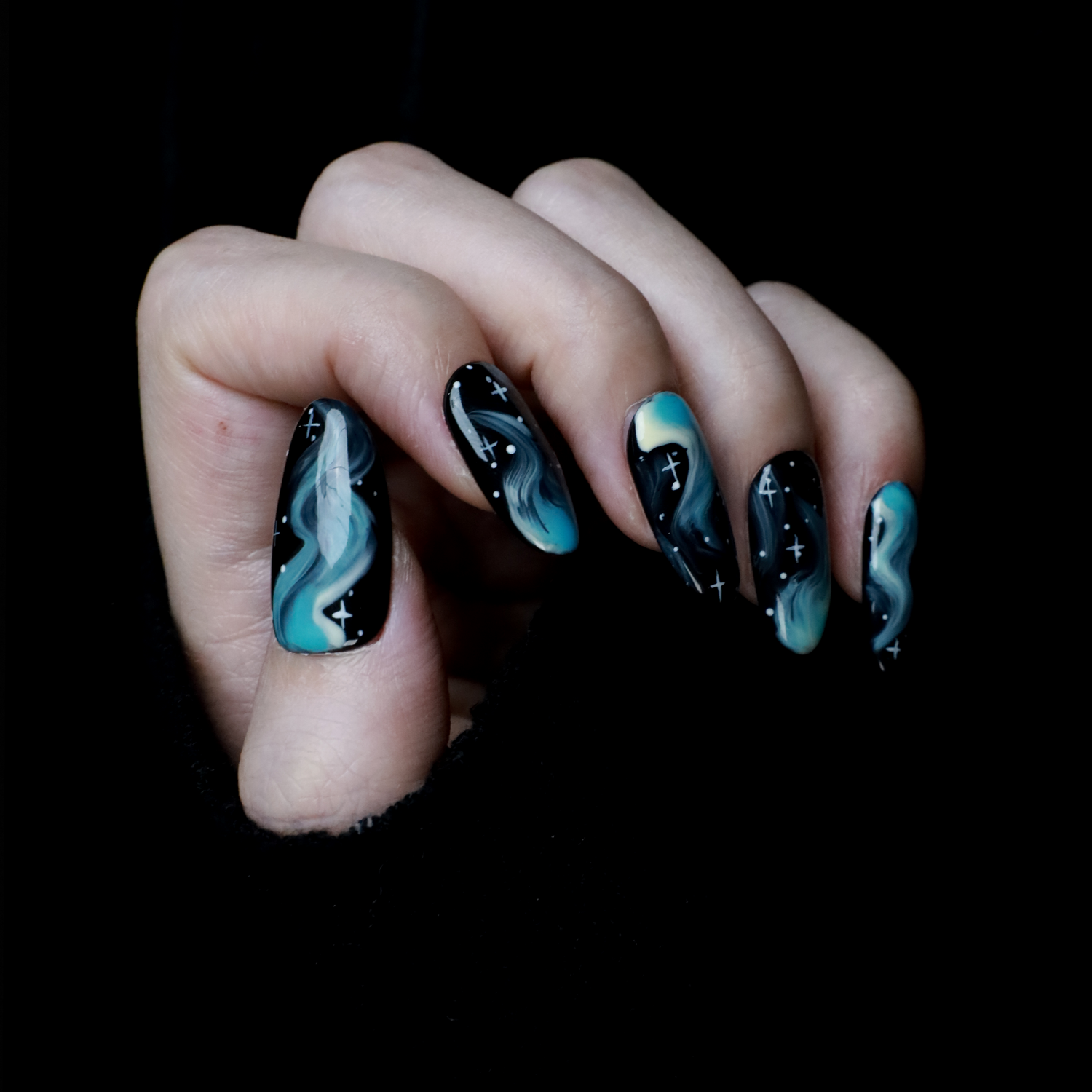 A hand showcases We Do Nails Amazing Blue Aurora Press-On Nails, featuring swirling blue and green patterns with white star-like dots on a black background, reminiscent of the northern lights. The hand is partially covered by a black sleeve.