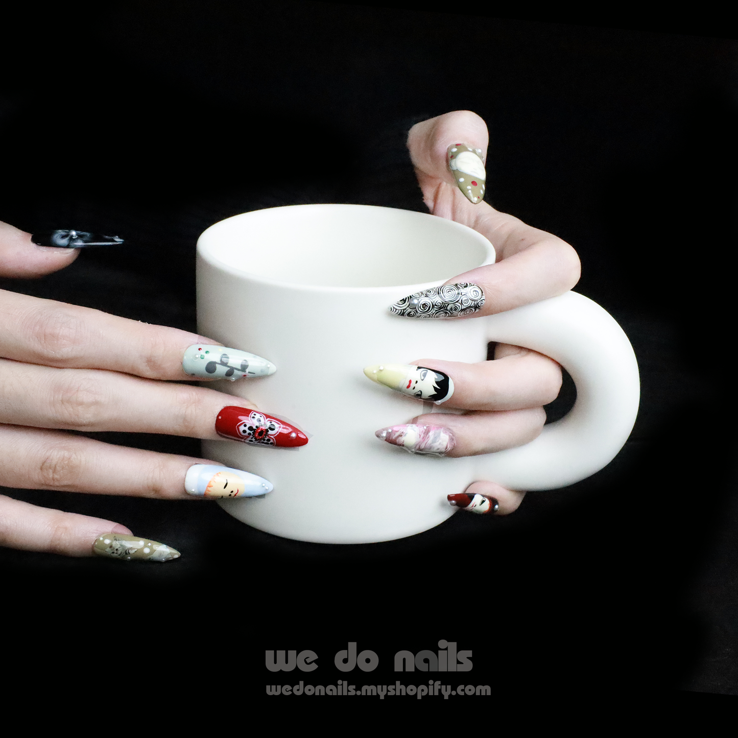 A hand with We Do Nails Cartoon Cuties Press-On Nails, featuring faces, abstract designs, and geometric shapes on a black background, holds a white teacup.