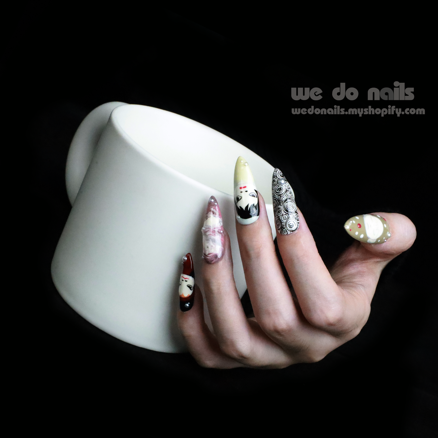 A hand with We Do Nails Cartoon Cuties Press-On Nails, featuring faces, abstract designs, and geometric shapes on a black background, holds a white teacup.