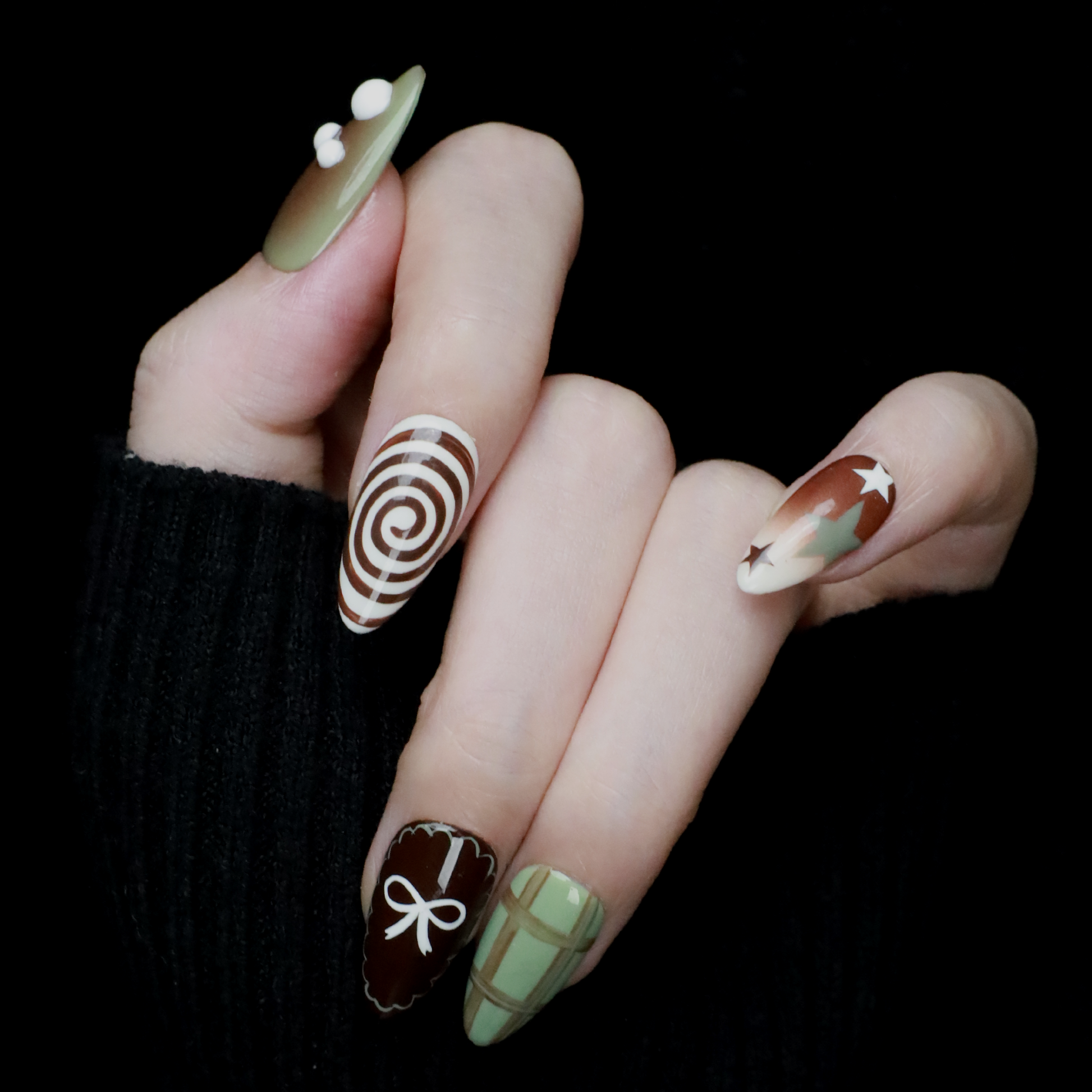 A hand displays the Retro Elegance Press-On Nails from We Do Nails, featuring artful designs: an olive green 3D pearl-accented nail, a spiral pattern, brown star-themed nails, a bow on brown, and green plaid. The classic elegance stands out against the black sweater.