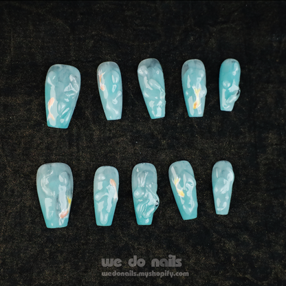 A hand featuring Fishy and 3D Water Press-Ons Nails in light blue holds a perfume bottle against a black background, highlighting the gorgeous nails and sleek design. We Do Nails with a website URL is displayed at the bottom.