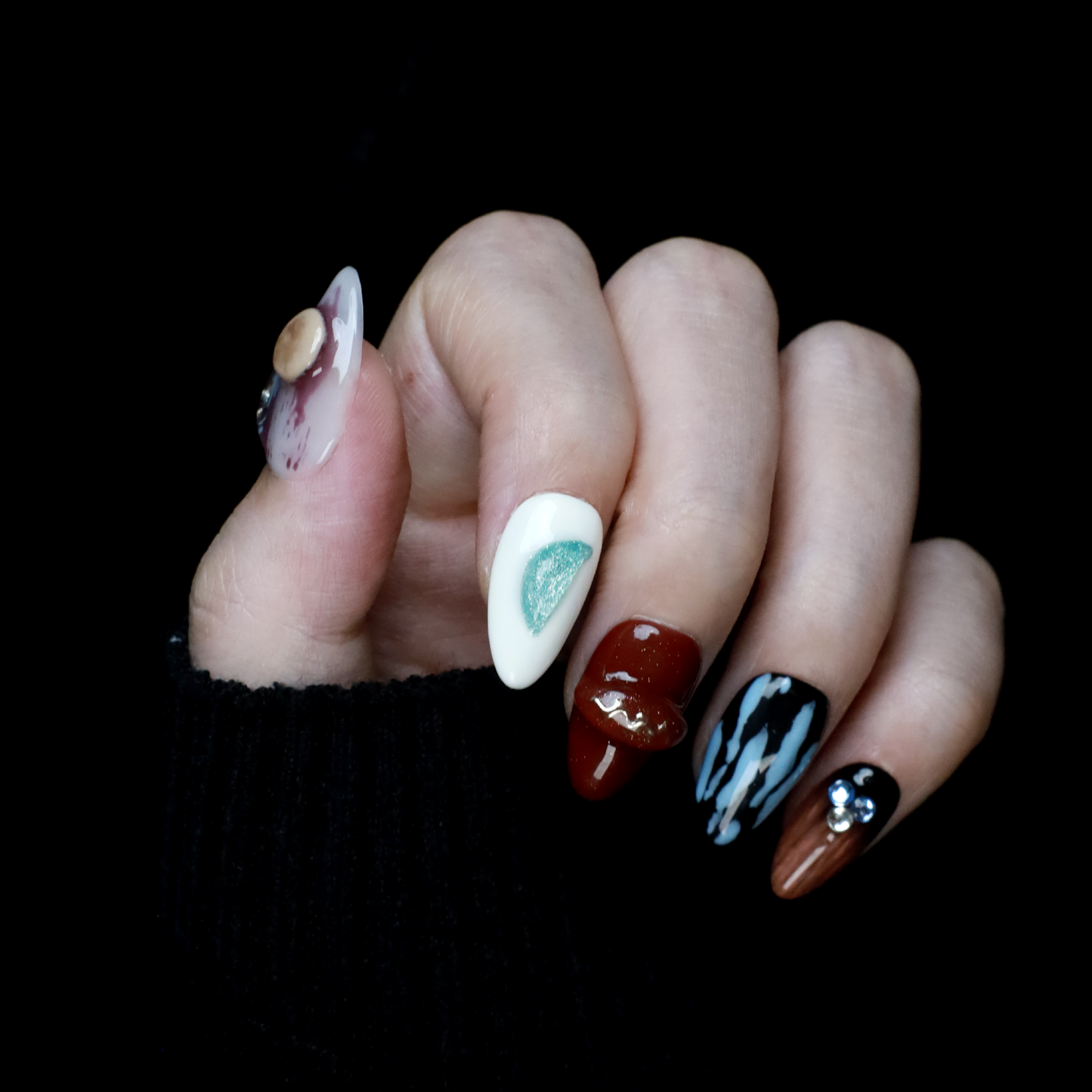 A hand elegantly displays Avant-Garde Luxe Press-On Nails by We Do Nails against a black backdrop. These nails, featuring 3D embellishments, boast white, maroon, and black hues with intricate patterns and metallic accents.