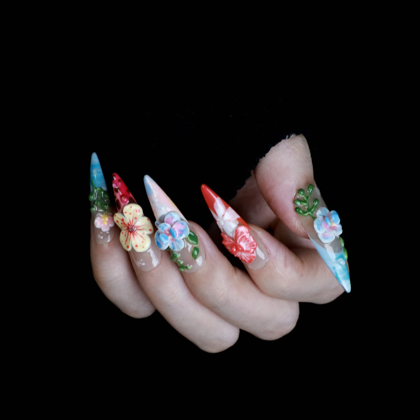 A hand shows off We Do Nails Floral Paradise Press-On Nails with long, pointed acrylics adorned with 3D flowers in shades of red, pink, yellow, and green. Set against a dark background, the vibrant blossoms truly stand out.