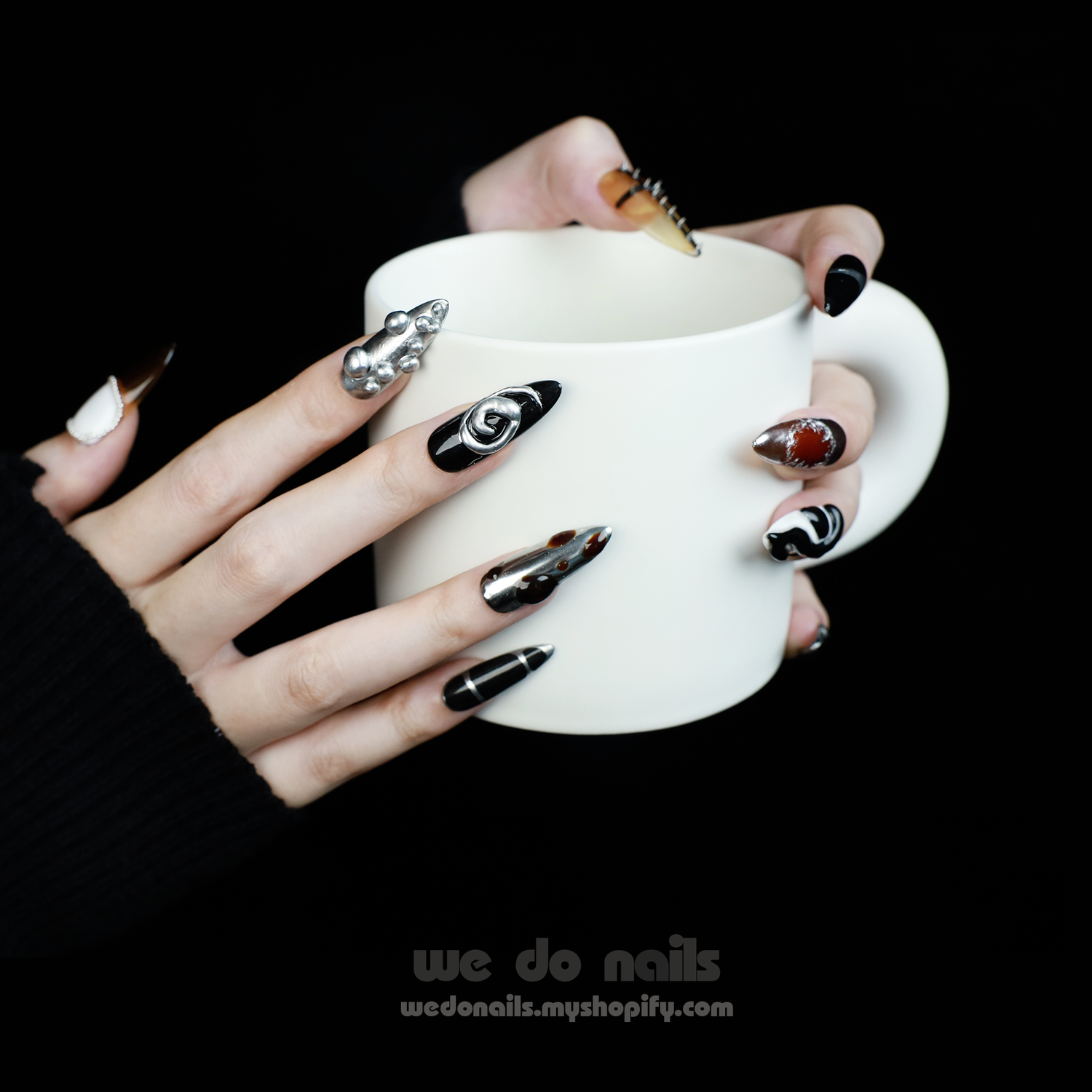 A hand holds a white mug, featuring long, intricately designed black and white Cool Rock Press-On Nails by We Do Nails from the Futuristic Noir Collection, adorned with metallic and gem accents against a dark background—an embodiment of avant-garde flair.