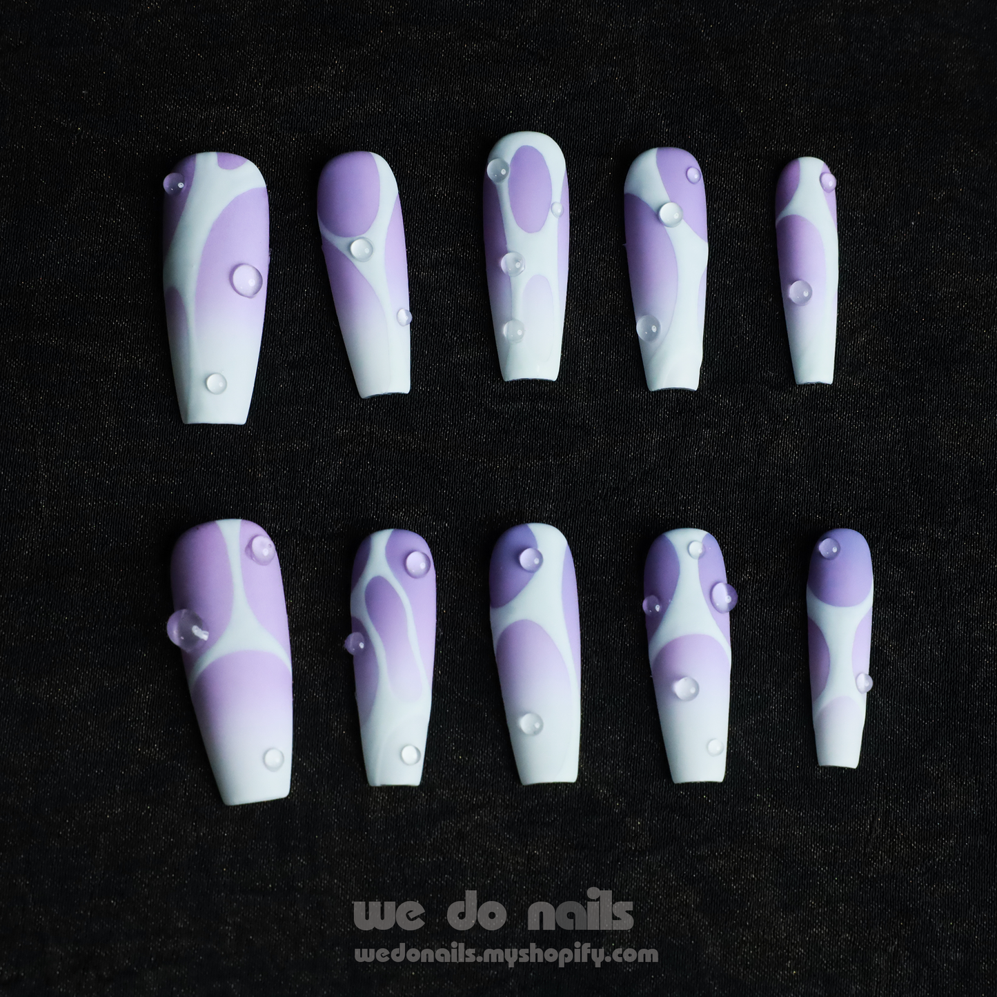 Holding a black mug, a person shows off Abstract Purple Press-On Nails by We Do Nails. These long, intricately designed nails feature teardrop patterns in a light purple to white gradient on a dark background. A website link is also included: WeDoNails.com.