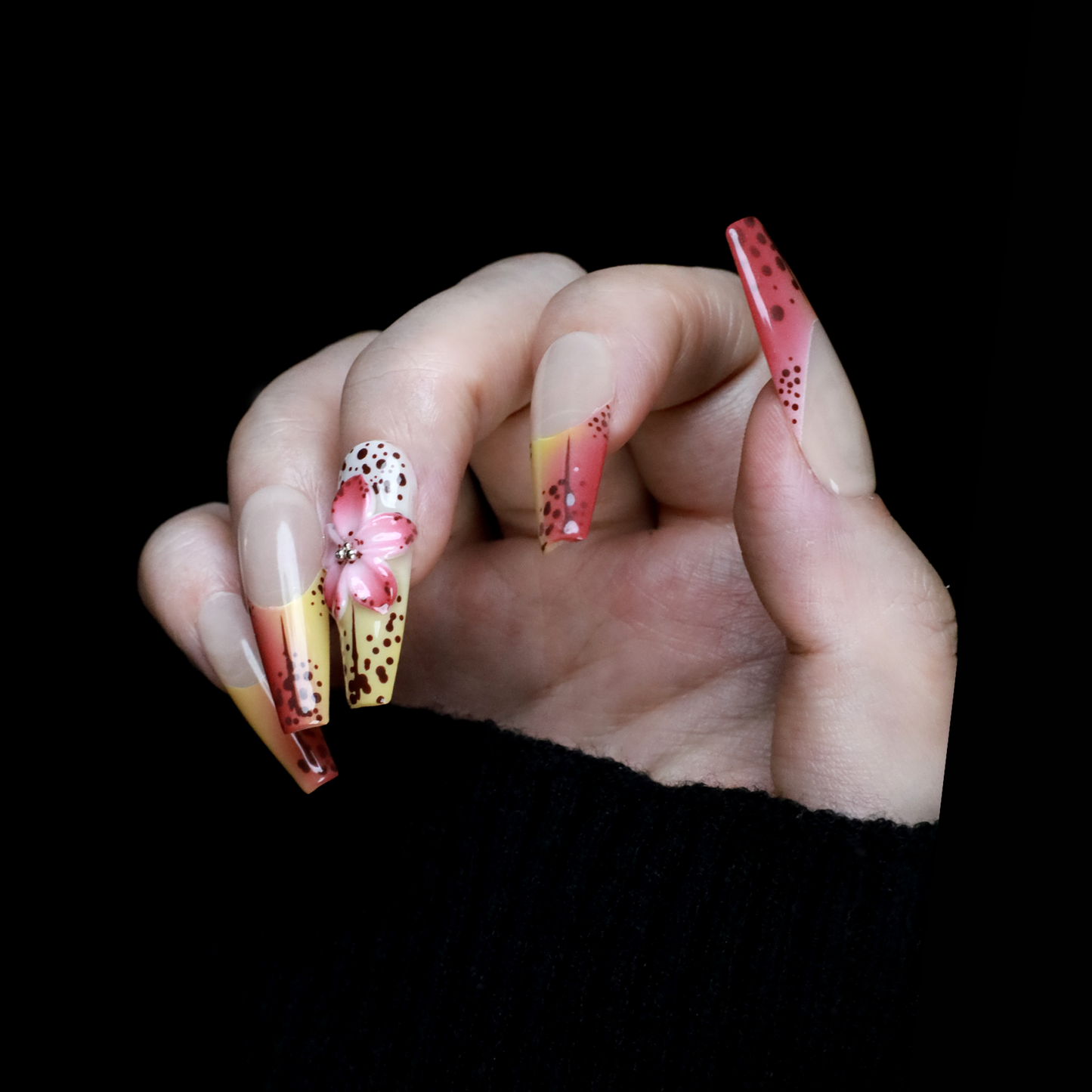 A hand with Sunset Bloom Press-On Nails by We Do Nails shows off long acrylics with a pink-yellow gradient and black dots. One has a 3D pink flower. Set against a black background, the hand is elegantly dressed in a black sleeve.