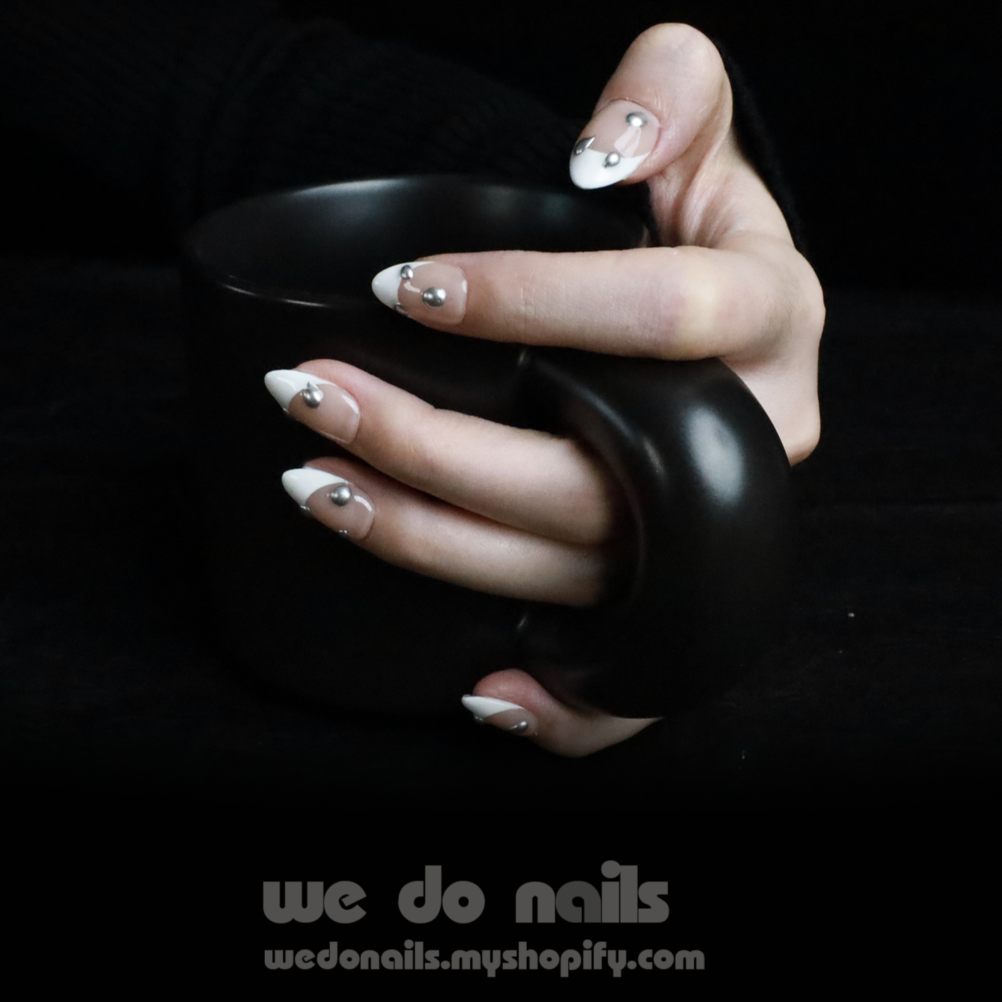 A hand with stylish, black and white Under the Moonlight French Tip Press On Nails adorned with small gems holds a matte black mug. Text at the bottom reads We Do Nails with a website link.