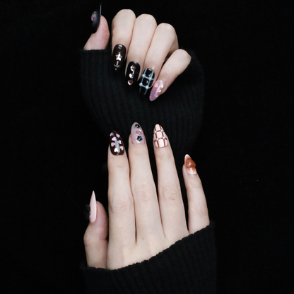 A hand with long, decorated nails appears against a black background, showcasing We Do Nails Midnight Mosaic Press-On Nails. The set includes various designs like pink hearts on clear, black polish with chain patterns, and sparkling gem accents with metallic stars on glossy black.