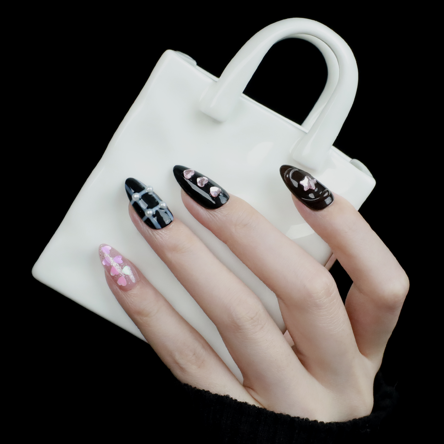 A hand with long, decorated nails appears against a black background, showcasing We Do Nails Midnight Mosaic Press-On Nails. The set includes various designs like pink hearts on clear, black polish with chain patterns, and sparkling gem accents with metallic stars on glossy black.