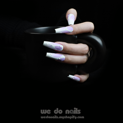 Holding a black mug, a person shows off Abstract Purple Press-On Nails by We Do Nails. These long, intricately designed nails feature teardrop patterns in a light purple to white gradient on a dark background. A website link is also included: WeDoNails.com.