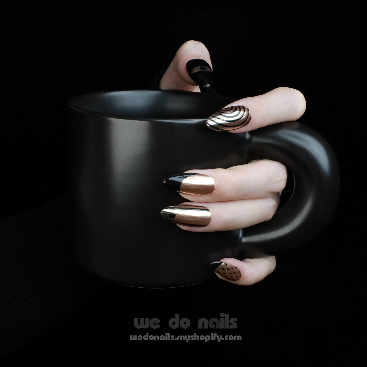 A hand with Egyptian-Inspired Press On Nails by We Do Nails holds a matte black mug against a dark background. The nails showcase metallic gold accents, striking tips, striped patterns, and abstract Eye of Horus elements.