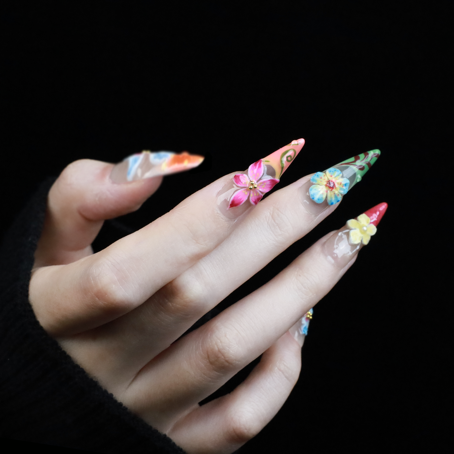 A hand shows off We Do Nails Floral Paradise Press-On Nails with long, pointed acrylics adorned with 3D flowers in shades of red, pink, yellow, and green. Set against a dark background, the vibrant blossoms truly stand out.