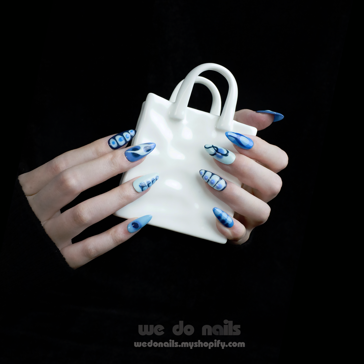 A person with Azure Waves Press-On Nails by We Do Nails, showcasing oceanic beauty and geometric designs, holds a small white handbag. The black background highlights the nails and bag, accentuating the nail arts blue waves.