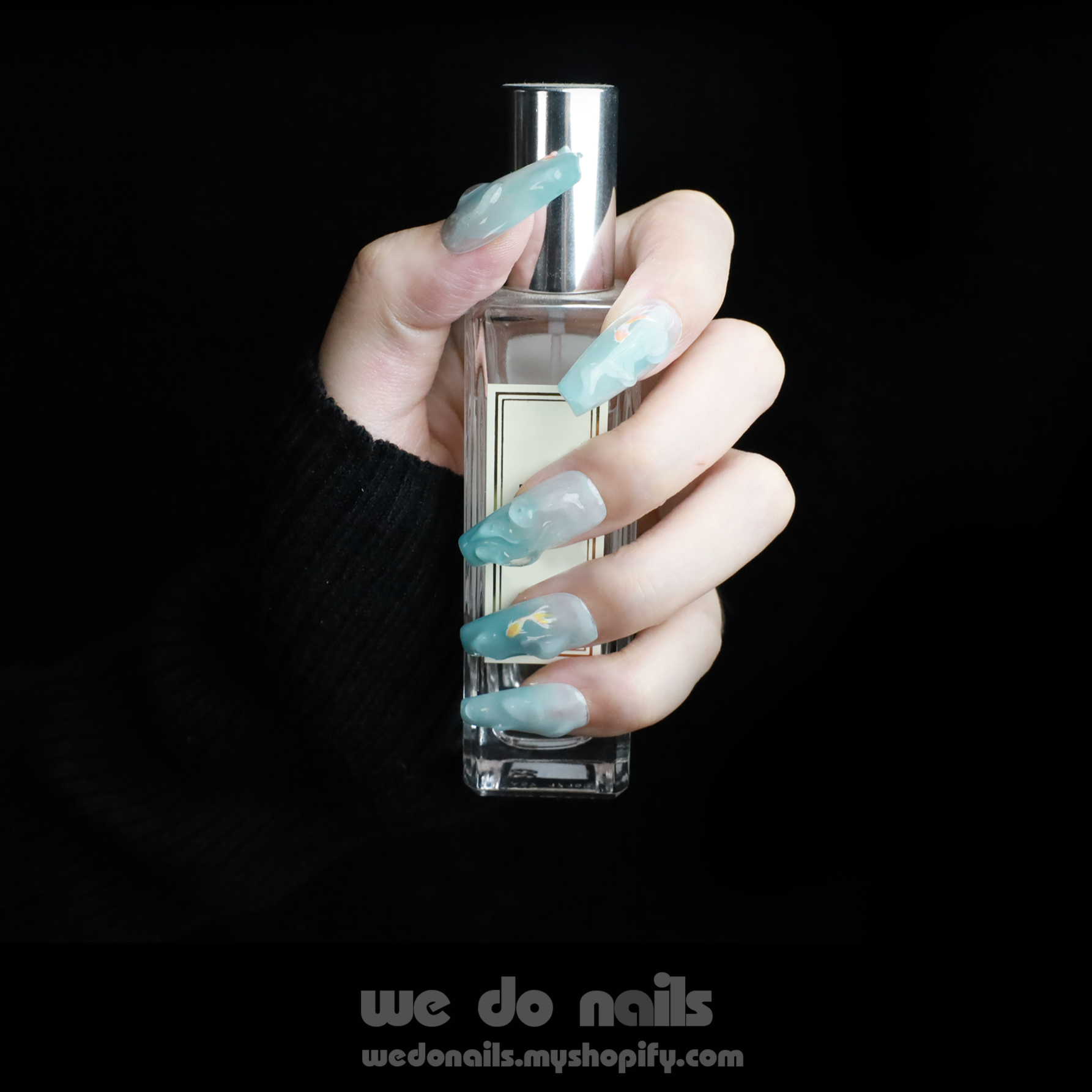 A hand featuring Fishy and 3D Water Press-Ons Nails in light blue holds a perfume bottle against a black background, highlighting the gorgeous nails and sleek design. We Do Nails with a website URL is displayed at the bottom.