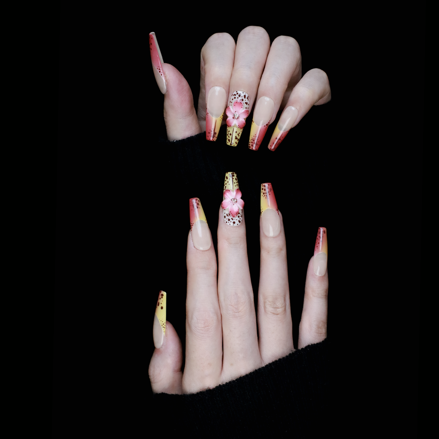 A hand with Sunset Bloom Press-On Nails by We Do Nails shows off long acrylics with a pink-yellow gradient and black dots. One has a 3D pink flower. Set against a black background, the hand is elegantly dressed in a black sleeve.