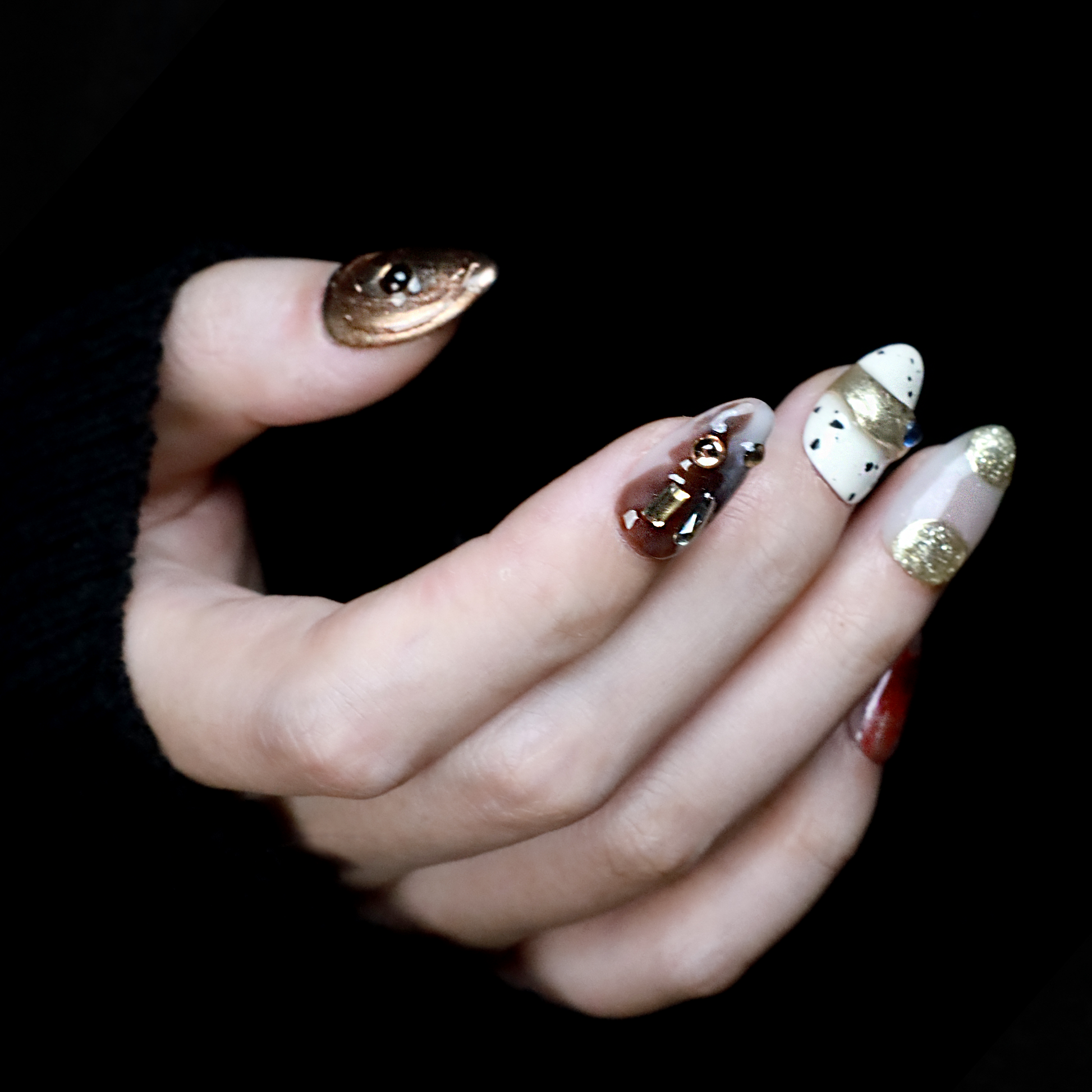 A hand elegantly displays Avant-Garde Luxe Press-On Nails by We Do Nails against a black backdrop. These nails, featuring 3D embellishments, boast white, maroon, and black hues with intricate patterns and metallic accents.