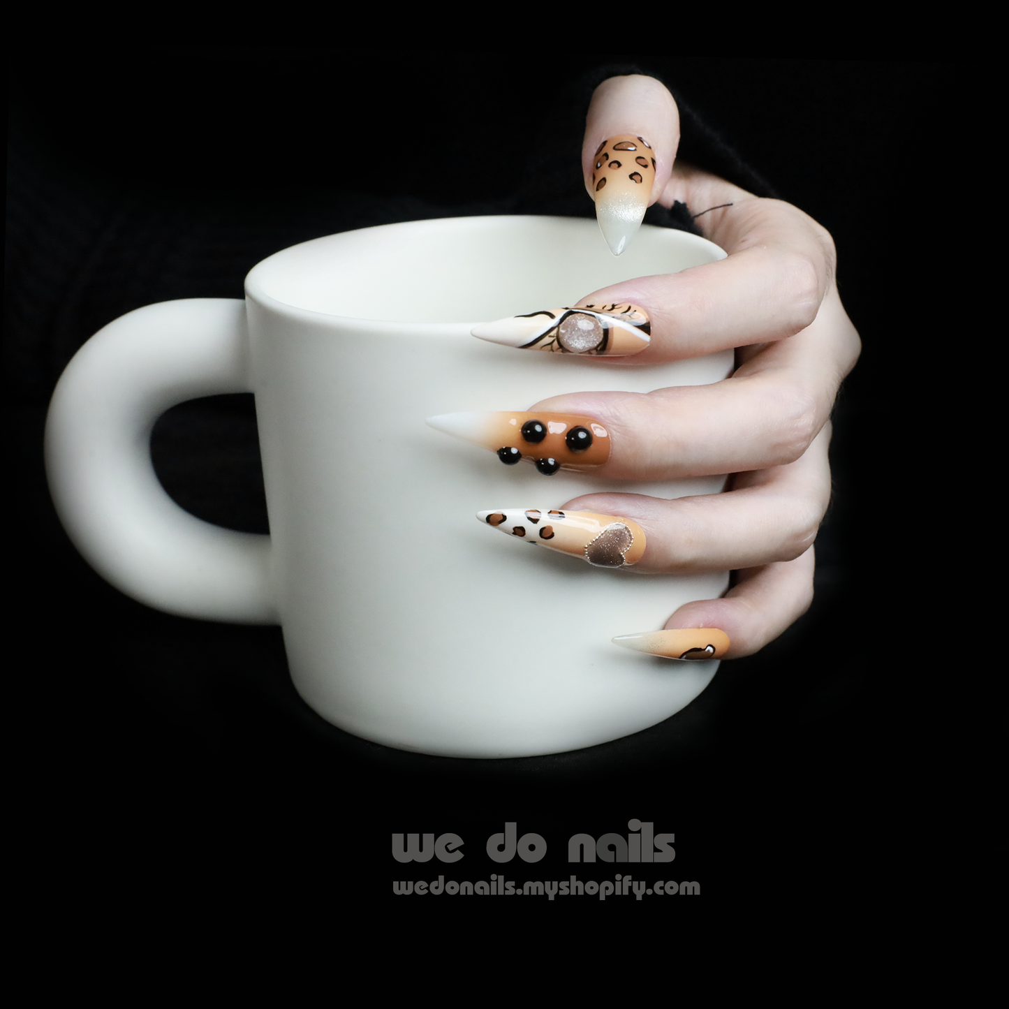 A hand with long, artistic nails featuring chic black and gold designs holds a white mug. The 3D Fierce Leopard Brown Press-On Nails by We Do Nails boast geometric shapes and rhinestones, embodying true Leopard Power.