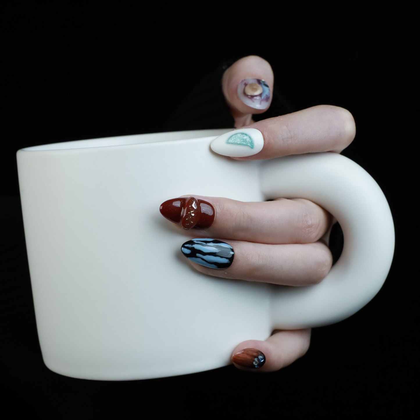 A hand elegantly displays Avant-Garde Luxe Press-On Nails by We Do Nails against a black backdrop. These nails, featuring 3D embellishments, boast white, maroon, and black hues with intricate patterns and metallic accents.