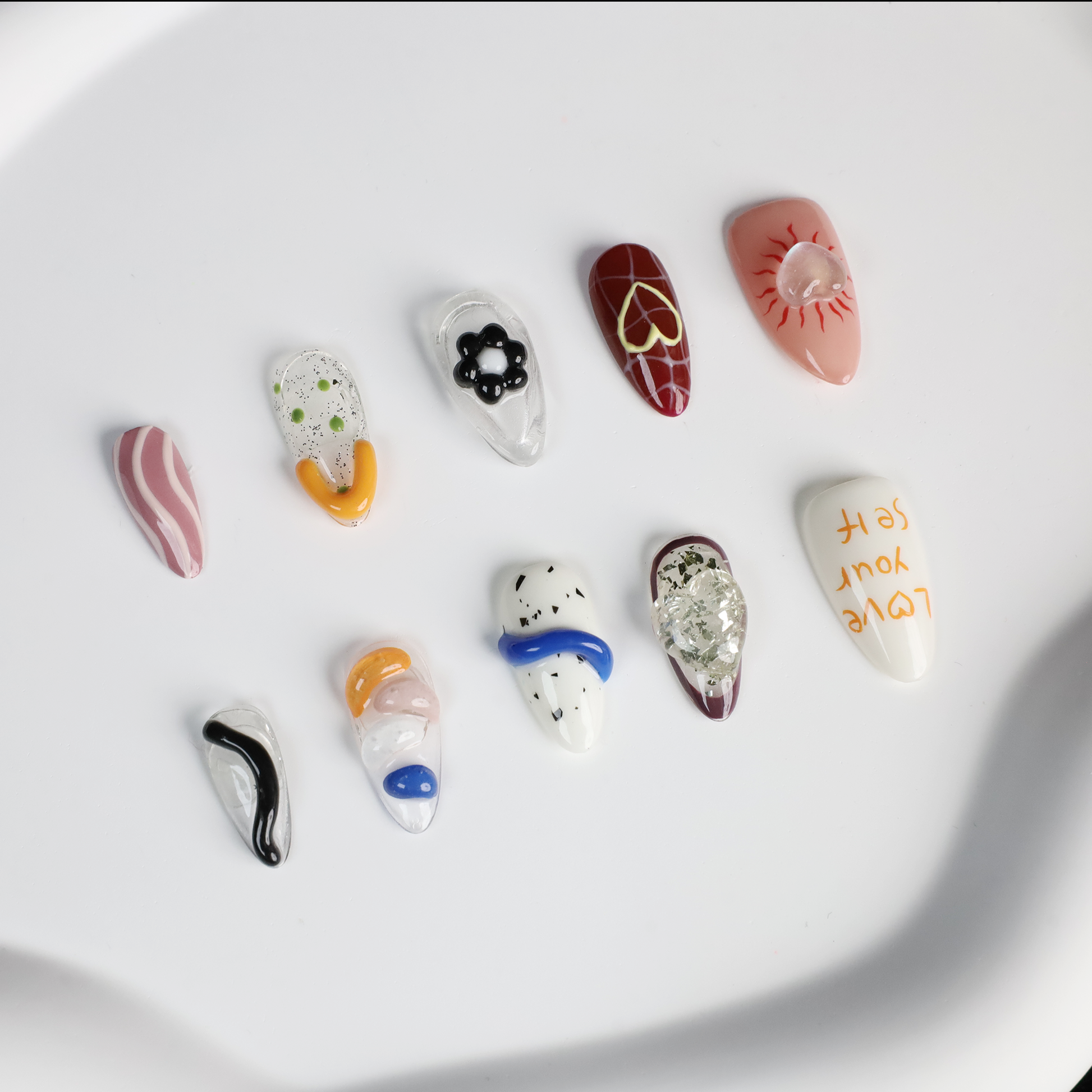 A hand displays vibrant Art-Inspired Abstract Press-On Nails by We Do Nails, set against a black backdrop. Each nail flaunts intricate hand-painted designs in black, orange, white, and blue with small accents and glitter. The wearer dons a long-sleeved black outfit.