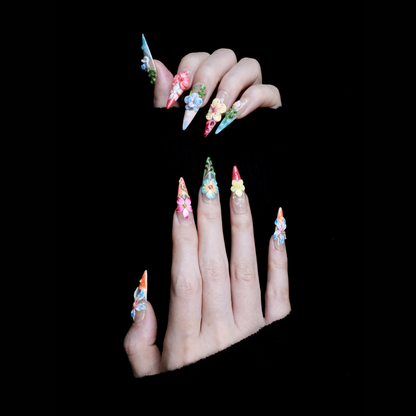 A hand shows off We Do Nails Floral Paradise Press-On Nails with long, pointed acrylics adorned with 3D flowers in shades of red, pink, yellow, and green. Set against a dark background, the vibrant blossoms truly stand out.