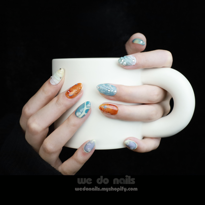 A persons hands hold a white mug, flaunting a colorful manicure from We Do Nails Ocean Breeze Press-On Nails. Each nail evokes a beach getaway with unique designs in blue, orange, and white adorned with intricate patterns and embellishments.