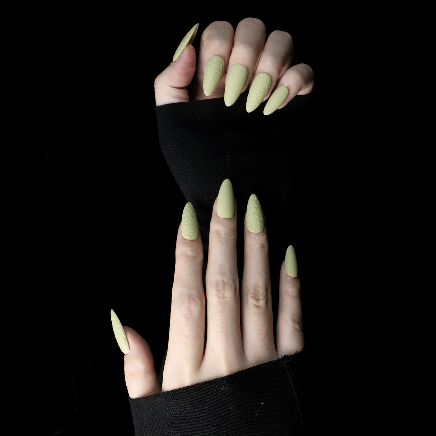 A person displays their crossed hands against a black background, showcasing We Do Nails Earthy Olive Green Press-On Nails. The textured olive design is elegantly highlighted by the sleeves of their black garment.