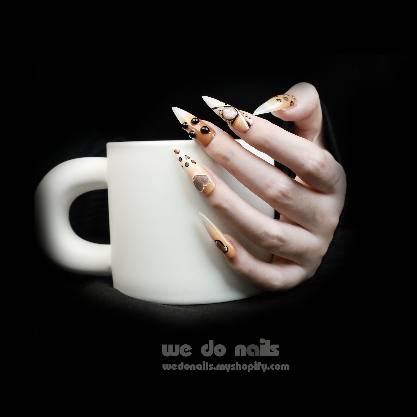 A hand with long, artistic nails featuring chic black and gold designs holds a white mug. The 3D Fierce Leopard Brown Press-On Nails by We Do Nails boast geometric shapes and rhinestones, embodying true Leopard Power.