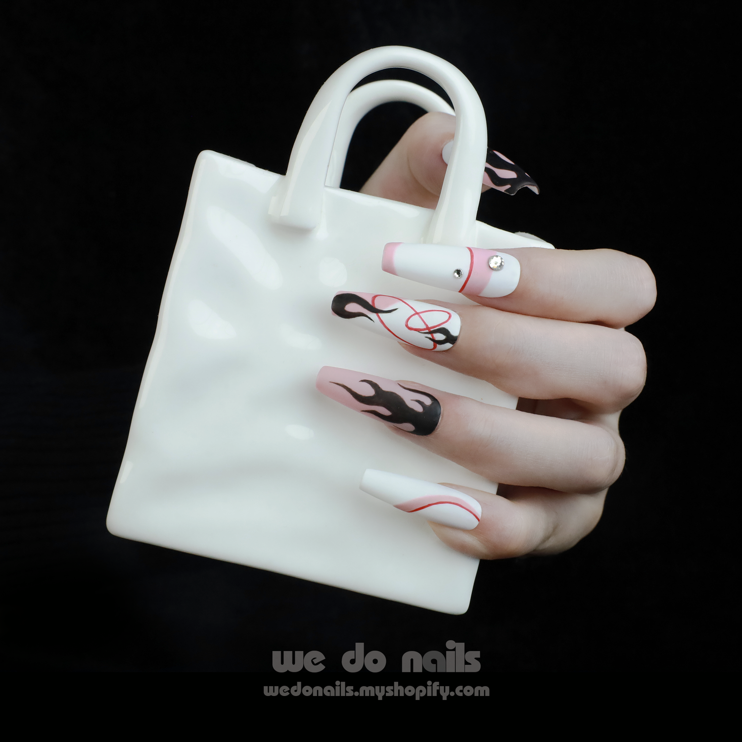 A hand with long, artistic nails from the We Do Nails Flame Passion Pink and Black Press-On Nails set features black and pink flame and minimalist designs, holding a small white handbag-shaped object against a dark background.