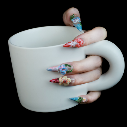 A hand shows off We Do Nails Floral Paradise Press-On Nails with long, pointed acrylics adorned with 3D flowers in shades of red, pink, yellow, and green. Set against a dark background, the vibrant blossoms truly stand out.