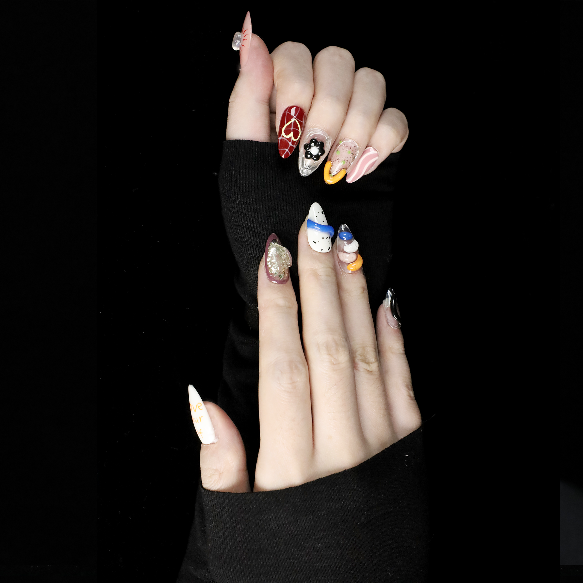 A hand displays vibrant Art-Inspired Abstract Press-On Nails by We Do Nails, set against a black backdrop. Each nail flaunts intricate hand-painted designs in black, orange, white, and blue with small accents and glitter. The wearer dons a long-sleeved black outfit.