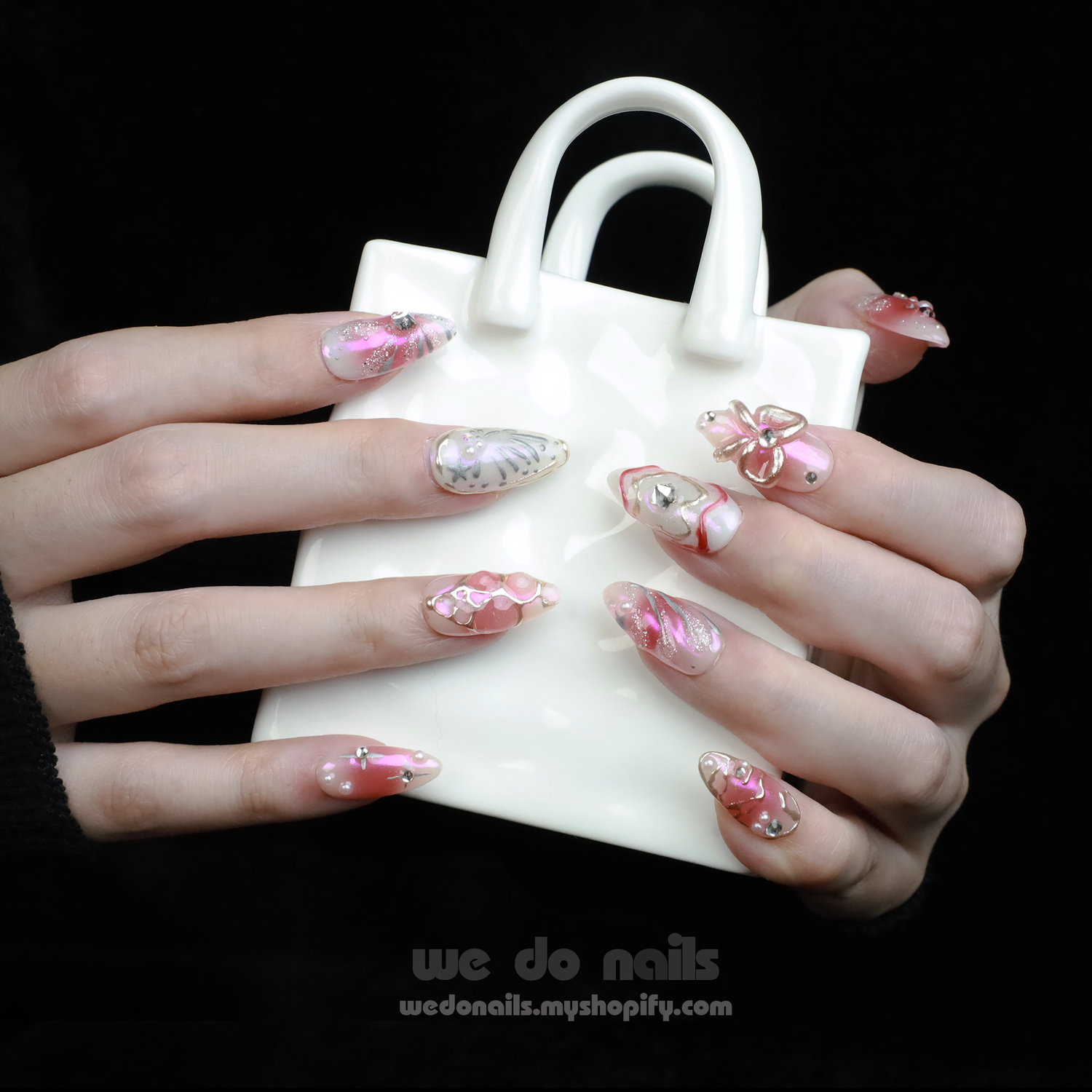 A person with intricately designed Blushing Charm Press-On Nails by We Do Nails holds a small, white ceramic handbag. The nails display soft pinks and abstract patterns in pink, white, and gold, accented with shimmering details and decorative embellishments.