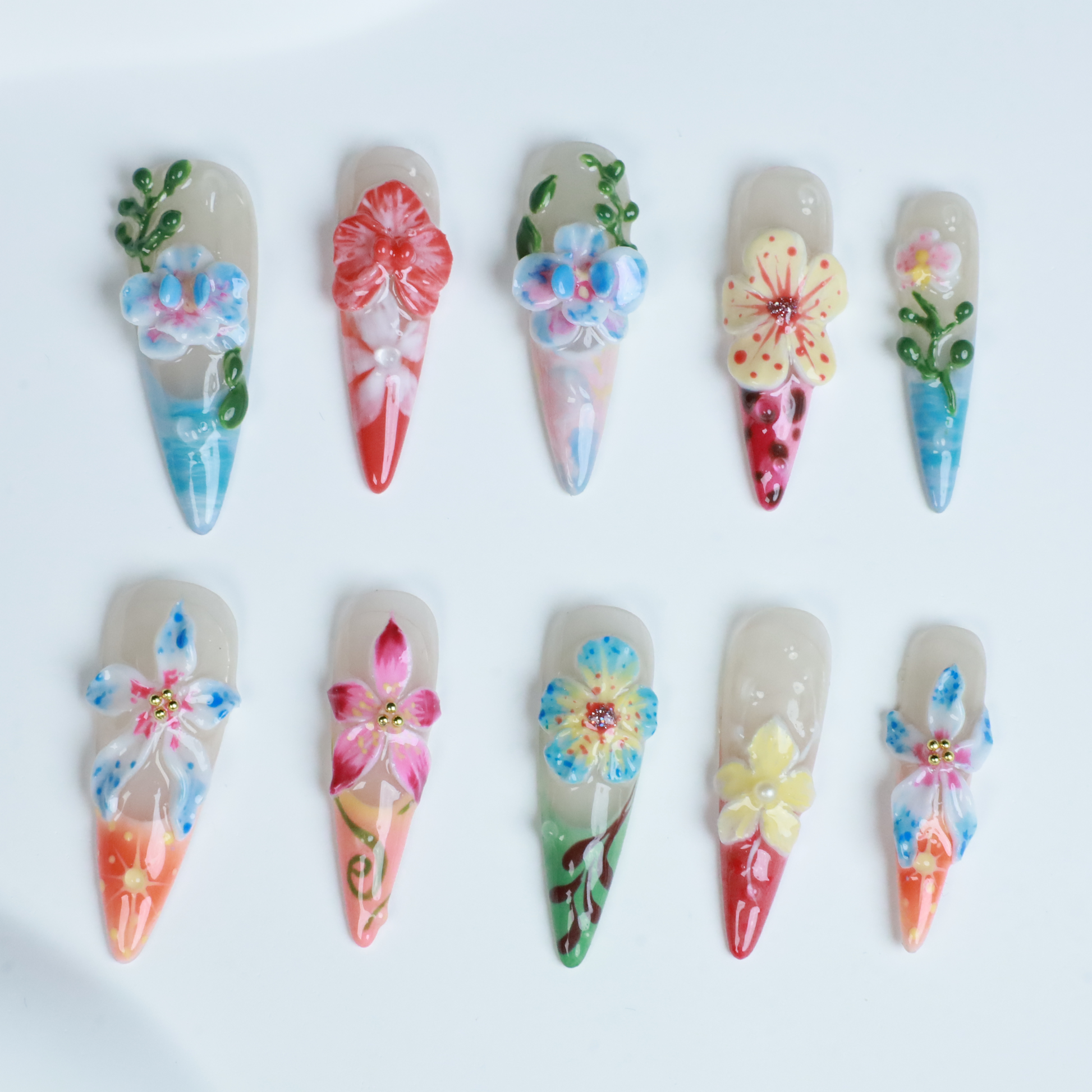 A hand shows off We Do Nails Floral Paradise Press-On Nails with long, pointed acrylics adorned with 3D flowers in shades of red, pink, yellow, and green. Set against a dark background, the vibrant blossoms truly stand out.