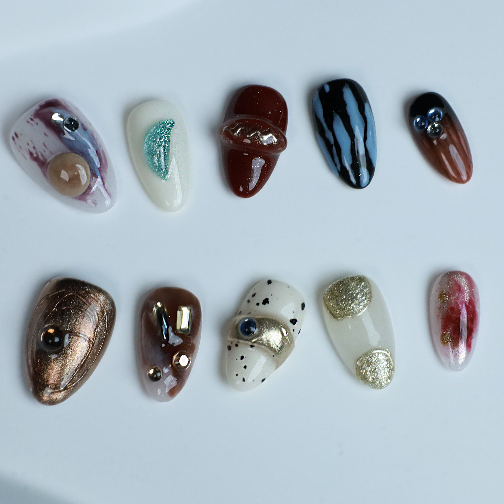 A hand elegantly displays Avant-Garde Luxe Press-On Nails by We Do Nails against a black backdrop. These nails, featuring 3D embellishments, boast white, maroon, and black hues with intricate patterns and metallic accents.