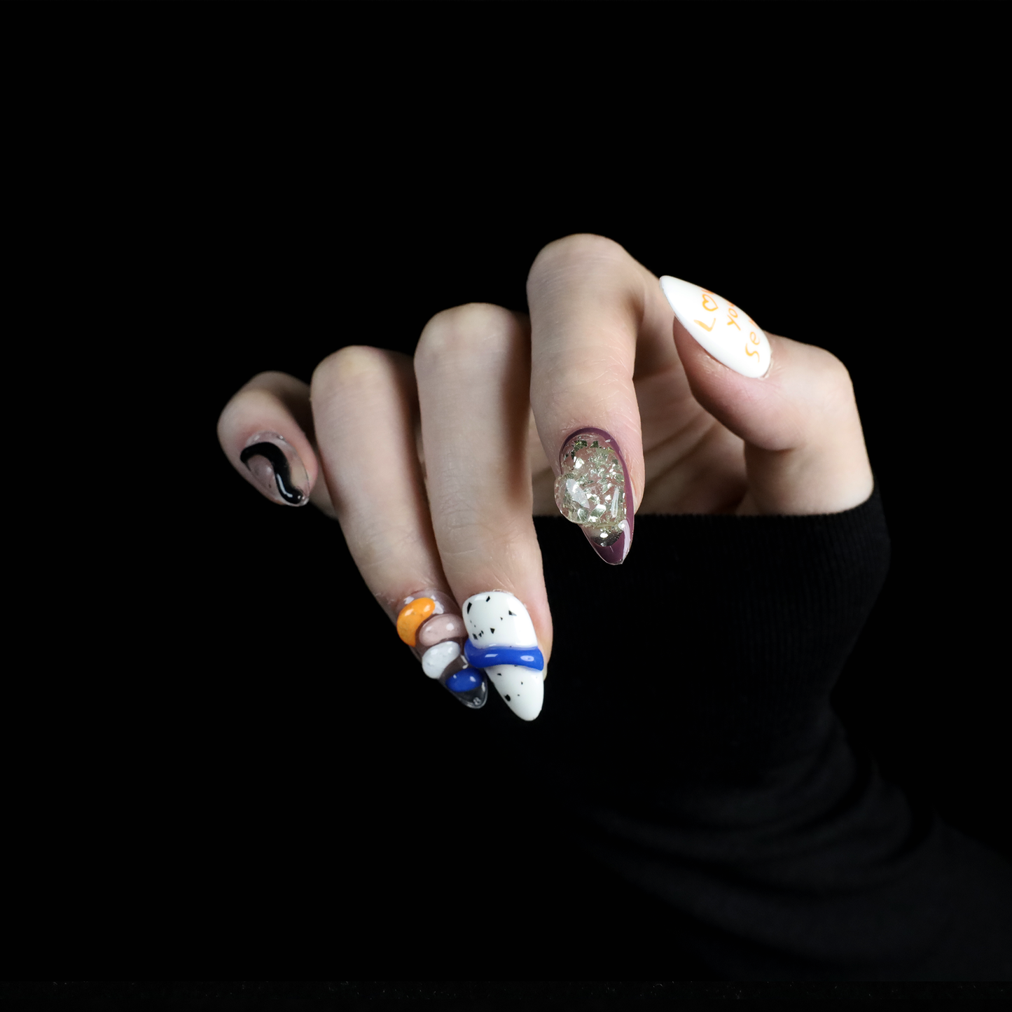 A hand displays vibrant Art-Inspired Abstract Press-On Nails by We Do Nails, set against a black backdrop. Each nail flaunts intricate hand-painted designs in black, orange, white, and blue with small accents and glitter. The wearer dons a long-sleeved black outfit.