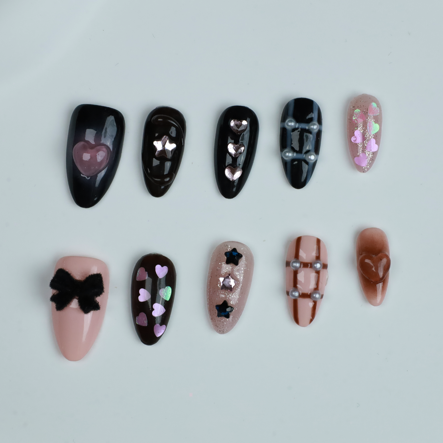 A hand with long, decorated nails appears against a black background, showcasing We Do Nails Midnight Mosaic Press-On Nails. The set includes various designs like pink hearts on clear, black polish with chain patterns, and sparkling gem accents with metallic stars on glossy black.