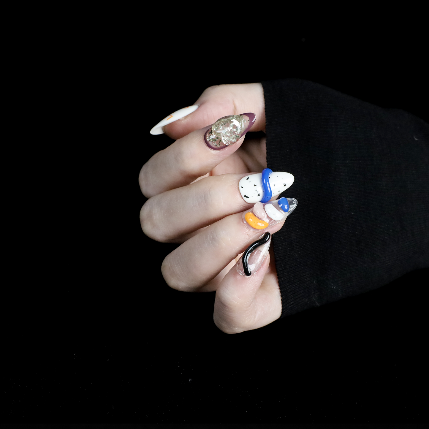 A hand displays vibrant Art-Inspired Abstract Press-On Nails by We Do Nails, set against a black backdrop. Each nail flaunts intricate hand-painted designs in black, orange, white, and blue with small accents and glitter. The wearer dons a long-sleeved black outfit.