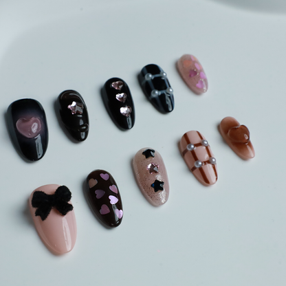 A hand with long, decorated nails appears against a black background, showcasing We Do Nails Midnight Mosaic Press-On Nails. The set includes various designs like pink hearts on clear, black polish with chain patterns, and sparkling gem accents with metallic stars on glossy black.