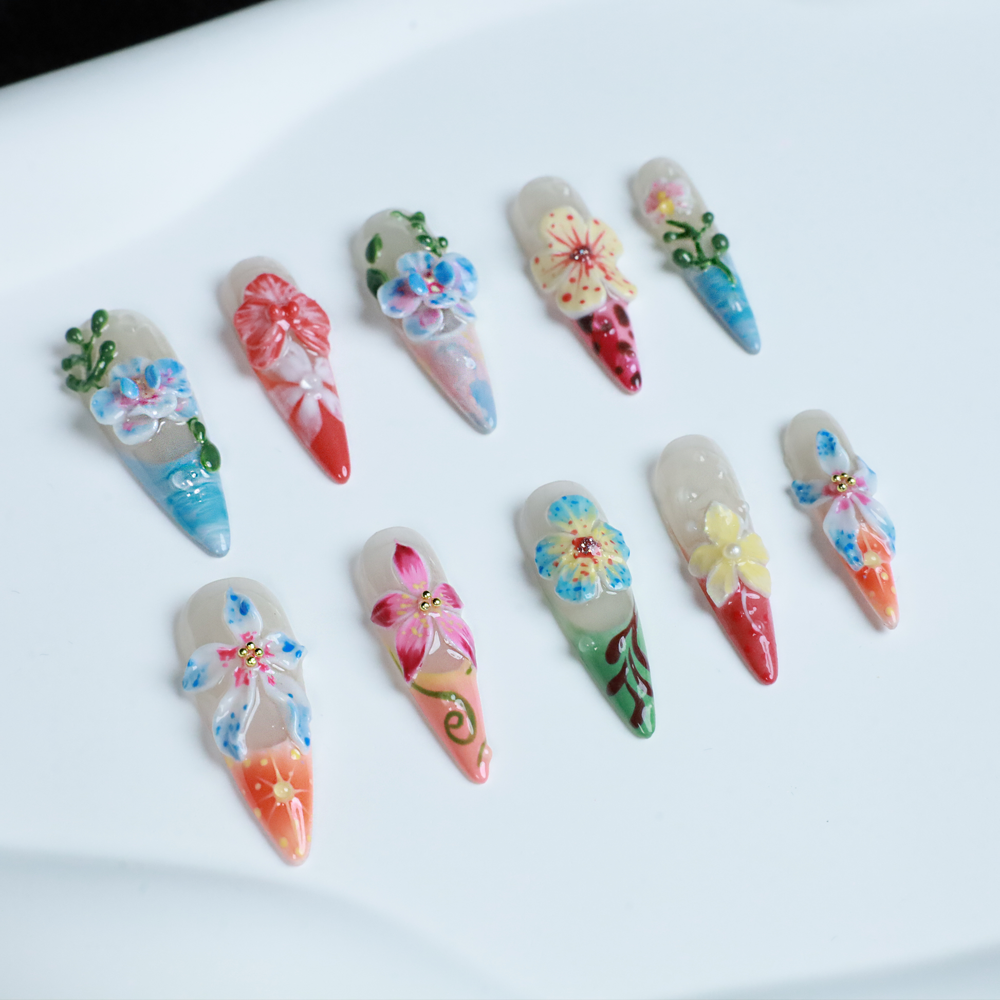 A hand shows off We Do Nails Floral Paradise Press-On Nails with long, pointed acrylics adorned with 3D flowers in shades of red, pink, yellow, and green. Set against a dark background, the vibrant blossoms truly stand out.