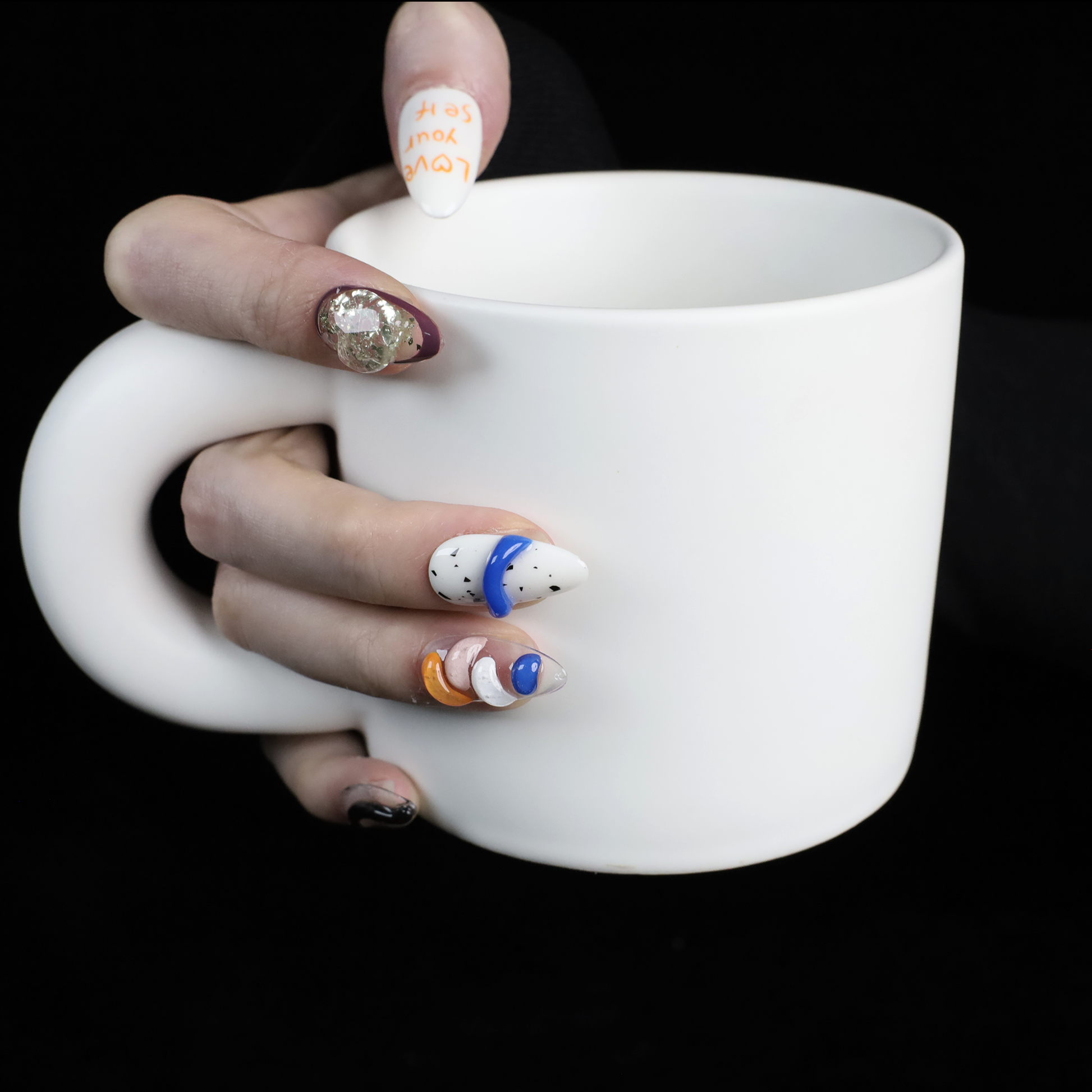 A hand displays vibrant Art-Inspired Abstract Press-On Nails by We Do Nails, set against a black backdrop. Each nail flaunts intricate hand-painted designs in black, orange, white, and blue with small accents and glitter. The wearer dons a long-sleeved black outfit.