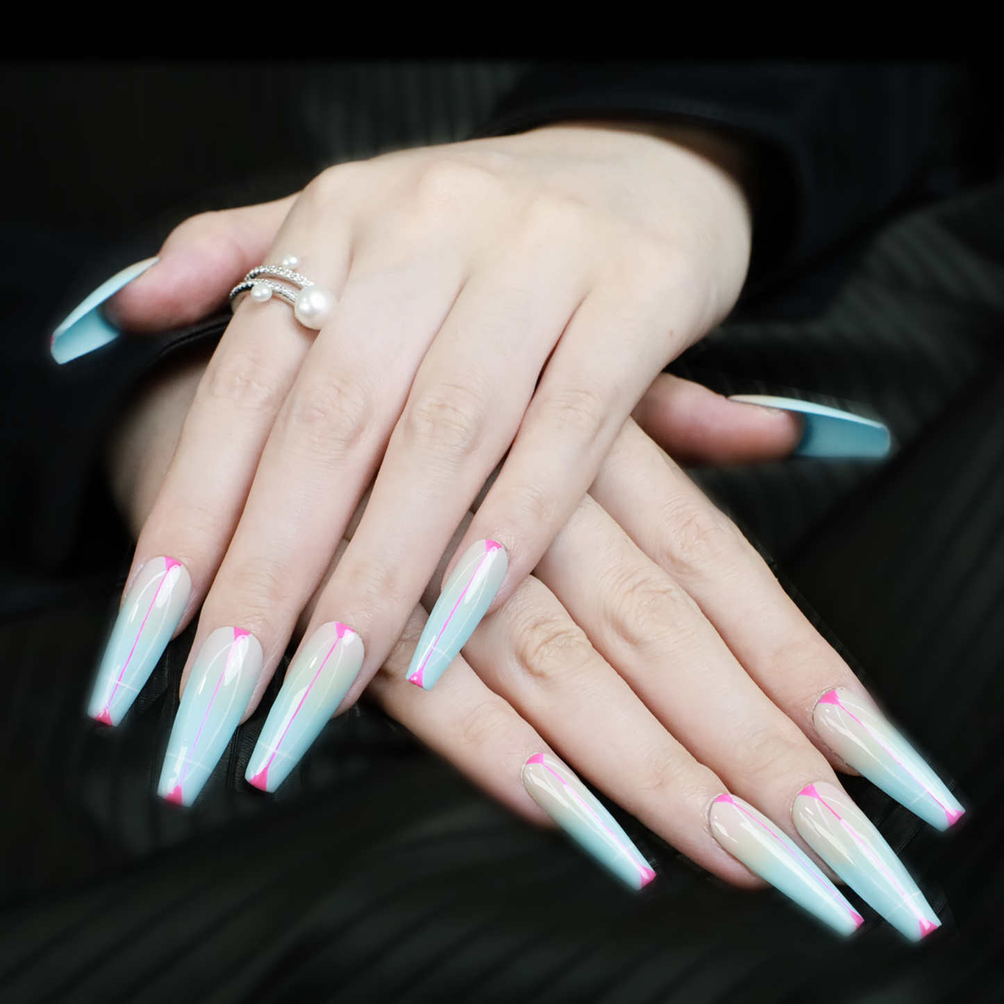 A pair of hands with long, pointed We Do Nails Sky Dream Press-On Nails features an iridescent light blue and pink design. A pearl ring on the index finger adds romantic charm, and the dark background highlights their beauty.